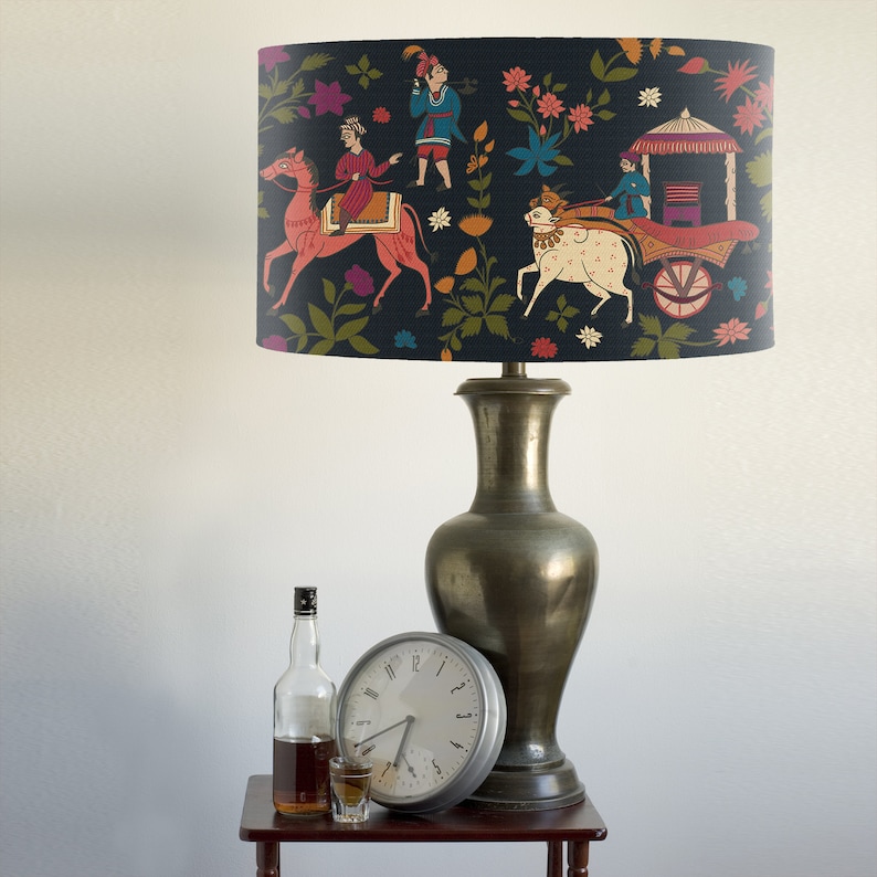 May include: A black lampshade with a colorful Indian-inspired design featuring people riding animals and a carriage. The lampshade is on a lamp with a gold-colored base.