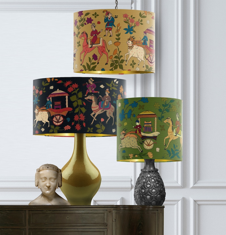May include: Three table lamps with different colored lampshades featuring a colorful Indian-inspired design of people riding horses and pulling a carriage. The lampshades are black, gold, and green.