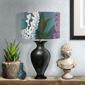 May include: A black ceramic table lamp with a floral patterned lampshade. The lampshade features white, pink, and green flowers on a blue background. The lamp is sitting on a wooden surface with a potted succulent plant and a small blue vase.