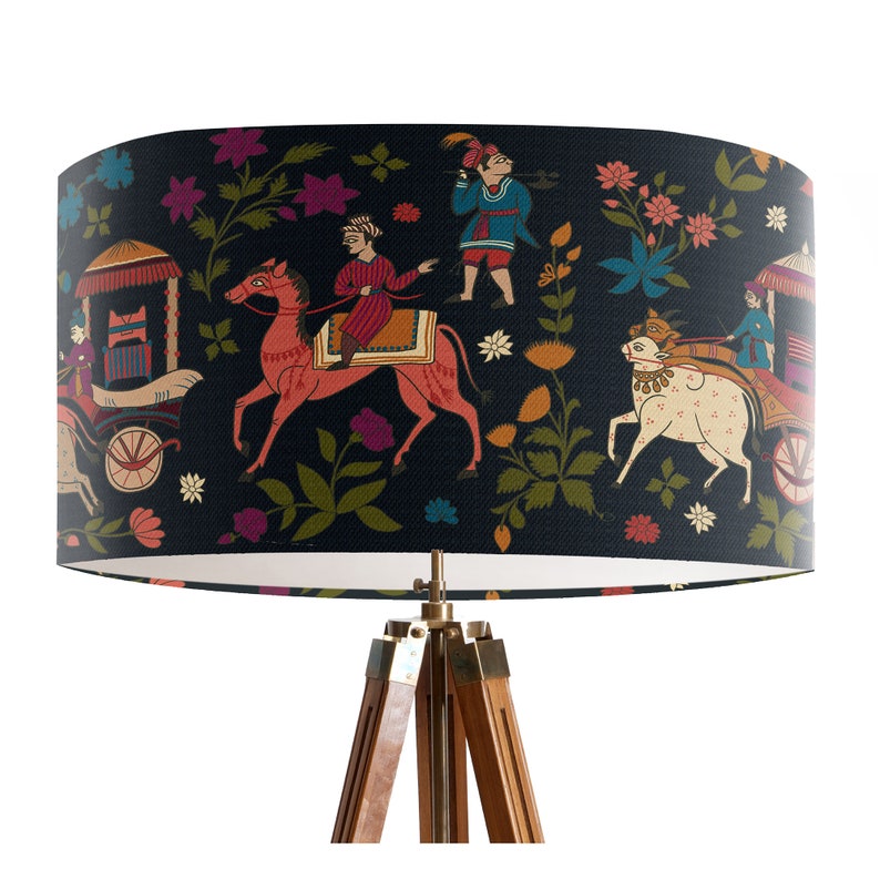 May include: A black lampshade with a colorful Indian-inspired design featuring people riding animals and floral patterns. The lampshade is on a wooden tripod stand.