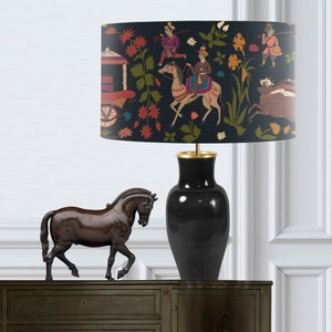 May include: A black lamp with a gold base and a lampshade featuring a colorful Indian-inspired design of people riding horses and a cart. A dark brown horse figurine is in the foreground.