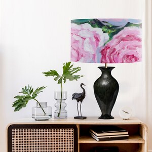K&ouml;nnte beinhalten: Eine schwarze Keramik-Tischlampe mit einem rosafarbenen Blumenlampenschirm. Die Lampe steht auf einem Holztisch mit einer schwarzen Kranichfigur, zwei klaren Glasvasen mit gr&uuml;nen Pflanzen und einer wei&szlig;en Uhr.