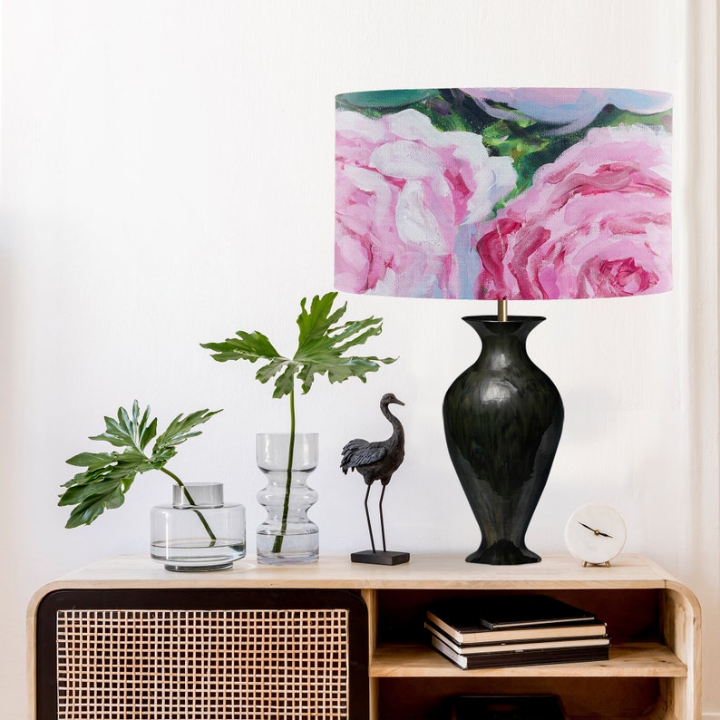 K&ouml;nnte beinhalten: Eine schwarze Keramik-Tischlampe mit einem rosafarbenen Blumenlampenschirm. Die Lampe steht auf einem Holztisch mit einer schwarzen Kranichfigur, zwei klaren Glasvasen mit gr&uuml;nen Pflanzen und einer wei&szlig;en Uhr.