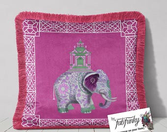 Op de afbeelding: Een magenta kussen met een gefranjerde rand en een witte geometrische rand. Het kussen heeft een gedetailleerde illustratie van een olifant versierd met bloemmotieven en een pagode op zijn rug.