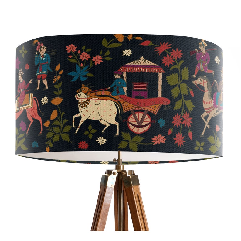 May include: A black lampshade with a colorful Indian-inspired design featuring people riding an ox-drawn cart, flowers, and trees. The lampshade is on a wooden tripod stand.