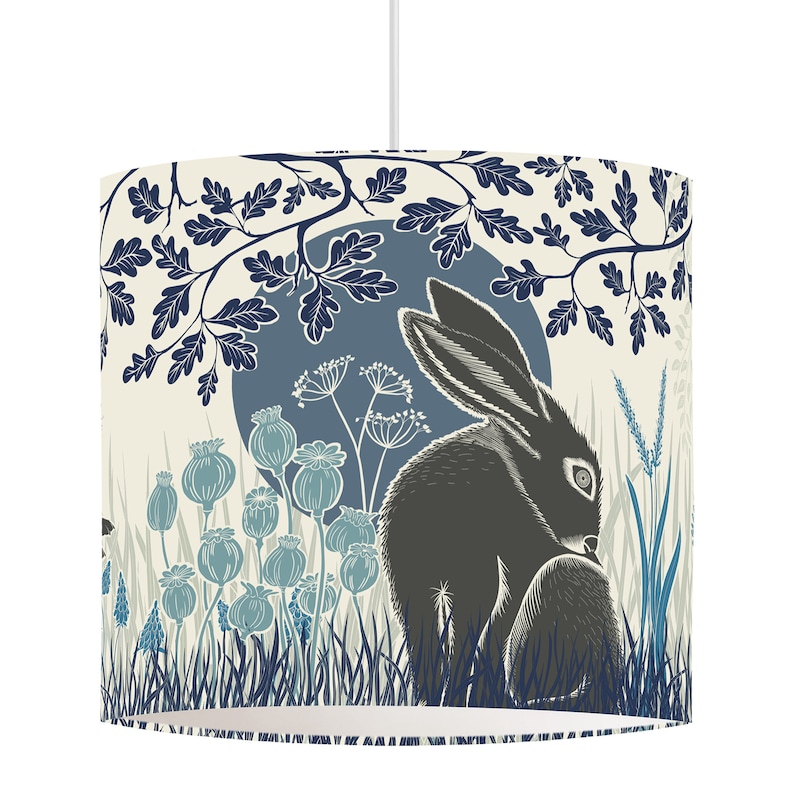 May include: A cream-colored lampshade with a blue and black illustration of a rabbit sitting in a field of flowers and grass under a blue moon. The design is stylized and features a repeating pattern of leaves and branches.