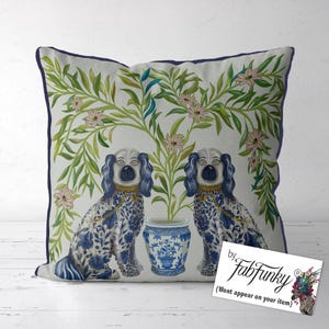 May include: Square cushion with a navy blue border. The design features two blue and white dog figures, a blue and white vase with a plant, and a background of green leaves and pink flowers. The cushion is square.