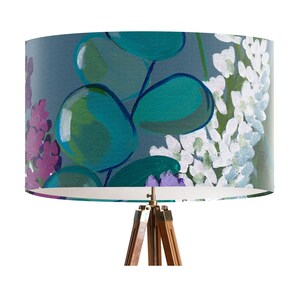 May include: A gray lampshade with a floral pattern featuring teal, green, purple, and white flowers. The lampshade is on a tripod stand with a wooden base.
