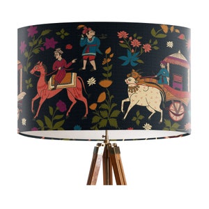 May include: A black lampshade with a white inner lining features a colorful Indian-inspired design with people riding animals and a bullock cart. The lampshade is on a tripod stand.