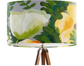 May include: A cylindrical lampshade featuring a floral design with large white and yellow flowers, green leaves, and a gray background. The lampshade is mounted on a wooden tripod lamp base with brass hardware. The overall aesthetic is artistic and decorative.