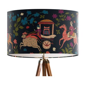 May include: A black lampshade with a white interior features a colorful Indian-inspired design with figures riding animals and a carriage. The lampshade is mounted on a wooden tripod stand.