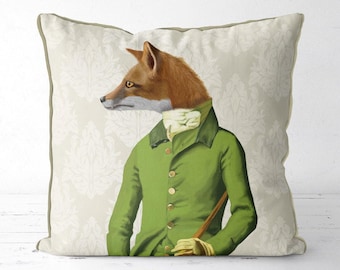 Op de afbeelding: Vierkantig sierkussen met een illustratie van een vos in een groene jas, witte cravat en wandelstok. De vos heeft bruine vacht en staat tegen een crèmekleurige achtergrond met een bloemenpatroon. De kussen heeft een lichtgroene rand.