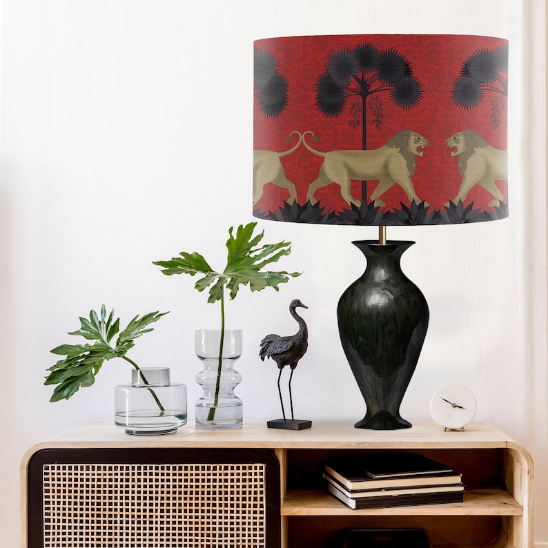 May include: A black lamp with a red lampshade featuring a repeating pattern of lions and palm trees. The lamp is on a wooden table with a black vase and a green plant.