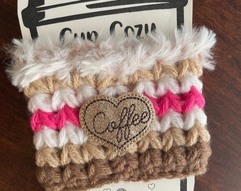 Könnte beinhalten: Ein gehäkeltes Kaffeetassen-Cozy mit einem braunen, weißen und rosafarbenen Streifenmuster. Das Cozy hat ein goldenes Glitzerherz mit dem Wort "Coffee" darauf. Das Cozy ist handgefertigt und hat einen weißen Kunstpelzbesatz.
