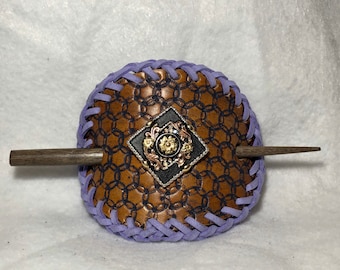 May include: A brown leather hair accessory with a chain pattern and a decorative metal embellishment. The accessory is edged with lavender stitching and includes a wooden stick for securing the hair. A unique hair accessory.