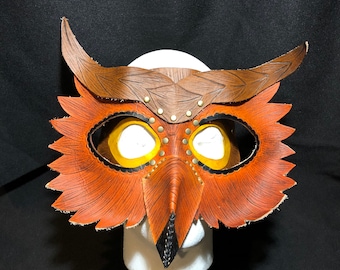 May include: A handcrafted leather owl mask with brown and orange hues. The mask features detailed feather-like edges, riveted accents, and a textured wooden-like appearance on the brow and horns. The eye openings are outlined in black and have yellow centers.