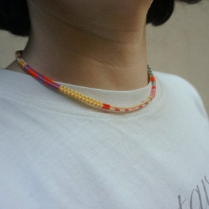 May include: A colorful necklace with a woven design. The necklace features a pattern of yellow, orange, pink, and purple threads. The necklace is worn around the neck and is visible against a white shirt.
