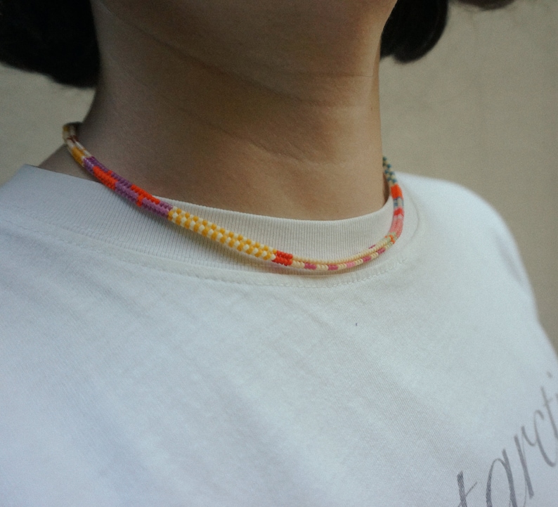 May include: A colorful necklace with a woven design. The necklace features a pattern of yellow, orange, pink, and purple threads. The necklace is worn around the neck and is visible against a white shirt.