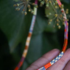 May include: Close-up of a colorful beaded bracelet with a variety of colors including orange, pink, blue, and white. The bracelet is held in a hand, with green foliage in the background. The beads are small and tightly strung.