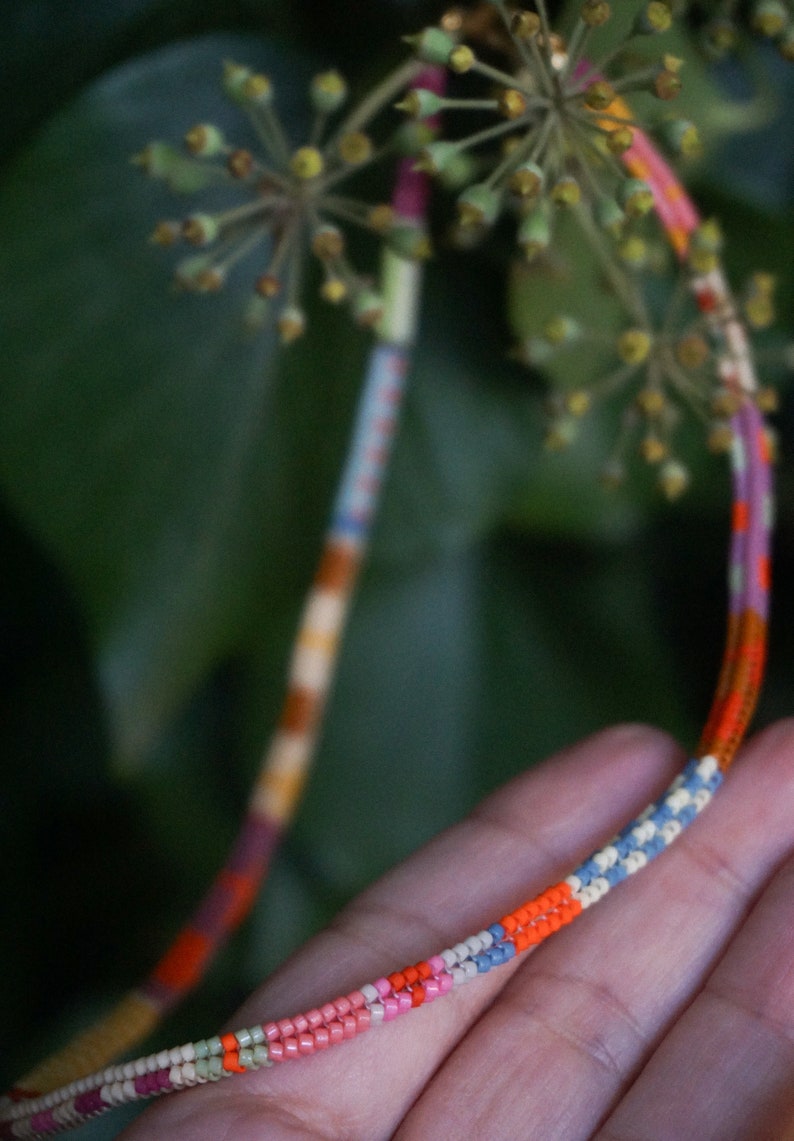 May include: Close-up of a colorful beaded bracelet with a variety of colors including orange, pink, blue, and white. The bracelet is held in a hand, with green foliage in the background. The beads are small and tightly strung.
