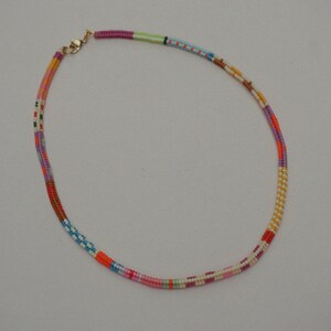 May include: A colorful beaded necklace with a gold-colored clasp. The necklace features a variety of colors, including pink, orange, purple, blue, and green. The beads are arranged in a striped pattern, creating a vibrant and eye-catching accessory.