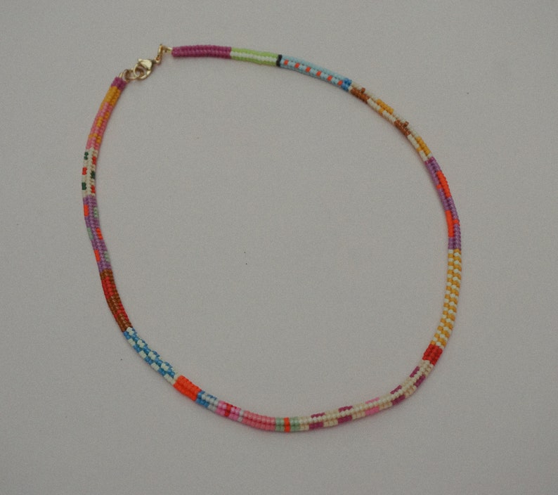 May include: A colorful beaded necklace with a gold-colored clasp. The necklace features a variety of colors, including pink, orange, purple, blue, and green. The beads are arranged in a striped pattern, creating a vibrant and eye-catching accessory.