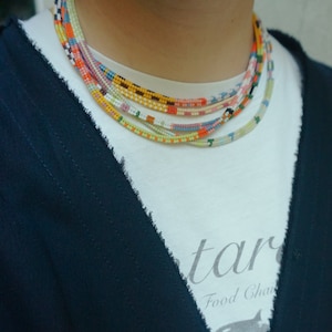 May include: A collection of colorful beaded necklaces in various patterns and colors, including orange, yellow, blue, and pink. The necklaces are layered and worn over a white t-shirt and a dark blue cardigan.