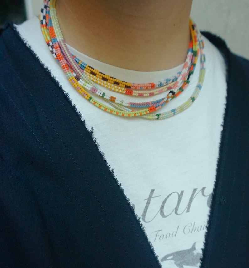 May include: A collection of colorful beaded necklaces in various patterns and colors, including orange, yellow, blue, and pink. The necklaces are layered and worn over a white t-shirt and a dark blue cardigan.
