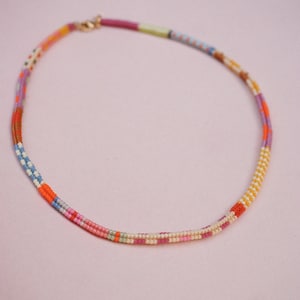 May include: A colorful beaded necklace with a gold clasp. The necklace features a variety of colors, including pink, orange, yellow, blue, and purple. The beads are arranged in a striped pattern, creating a vibrant and eye-catching accessory.
