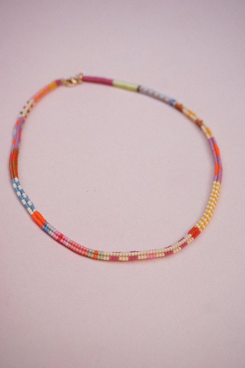 May include: A colorful beaded necklace with a gold clasp. The necklace features a variety of colors, including pink, orange, yellow, blue, and purple. The beads are arranged in a striped pattern, creating a vibrant and eye-catching accessory.