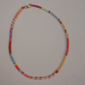 May include: A colorful beaded necklace with a gold-colored clasp. The necklace features a variety of colors, including pink, orange, yellow, green, and blue. The beads are arranged in a linear pattern, creating a vibrant and eye-catching accessory.
