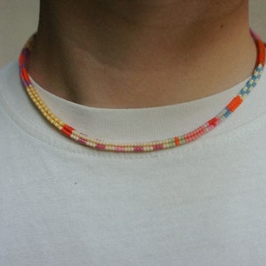 May include: A colorful beaded necklace with a repeating pattern of orange, yellow, pink, and blue beads. The necklace is worn around the neck and is against a white background.