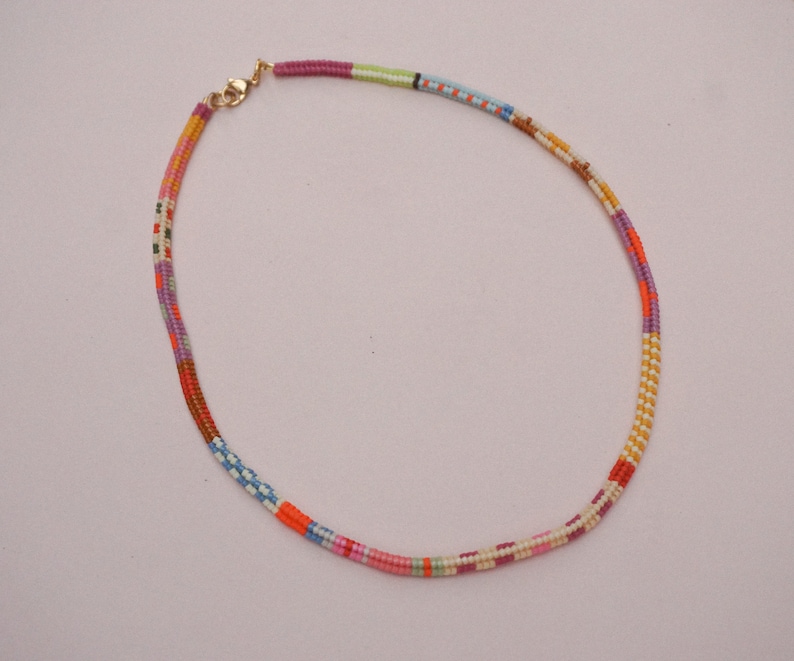 May include: A colorful beaded necklace with a gold-colored clasp. The necklace features a variety of colors, including red, orange, pink, purple, green, and blue. The beads are arranged in a linear pattern, creating a vibrant and eye-catching accessory.