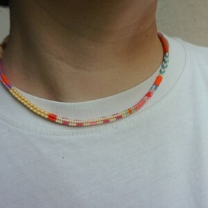 May include: A colorful beaded necklace with a striped pattern. The necklace features a variety of colors, including orange, yellow, pink, and blue. The necklace is worn around the neck and is displayed against a white t-shirt.