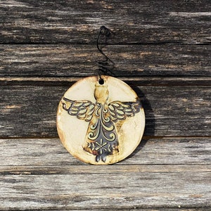 May include: A ceramic ornament shaped like an angel with a brown glaze and black details. The angel has wings and a halo. The ornament is hanging from a black wire.