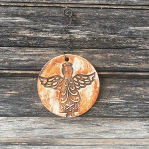 May include: A round, white ceramic ornament with a brown angel design. The angel has wings, a halo, and a flowing gown. The ornament is hanging from a wire loop.