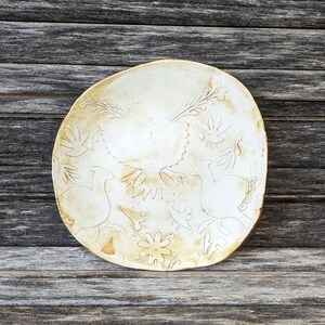 May include: A white ceramic plate with a raised design of stylized birds and flowers. The plate has a slightly off-white glaze and a rustic, handmade look.