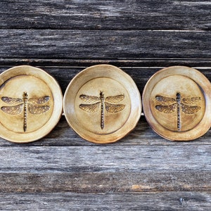 May include: Three small ceramic plates with a dragonfly design. The plates are a light brown color with a darker brown dragonfly design.