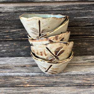 May include: A stack of four small, hand-formed ceramic bowls with an off-white, speckled glaze. Each bowl features a dark brown, etched dragonfly design on the exterior. The rims are uneven and wavy, with a light blue interior glaze visible.