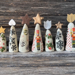 May include: A collection of seven ceramic Christmas tree figurines with various designs. Each tree is topped with a star, heart, or tulip. The trees feature snowflakes, holly, a nutcracker, and the text "Dear Heart".