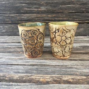May include: Two handmade ceramic tumblers with textured designs. One features a pattern of swirling roses, the other a floral motif. Both have a light brown exterior, a green interior, and sit on a wooden surface.