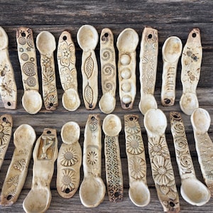 May include: A collection of handcrafted ceramic spoons with various embossed designs, including flowers, leaves, and geometric patterns. The spoons are beige and arranged on a weathered wooden surface, showcasing their unique textures and details.