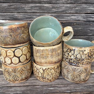 May include: A collection of six ceramic mugs with handles, each featuring a unique tan-colored exterior with embossed patterns including swirls, circles, and floral designs. The interior of each mug is glazed in a smooth, light sage green.