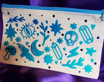 May include: A white canvas zippered pouch with a blue zipper. The pouch is decorated with a variety of blue, glittery, and iridescent designs, including stars, moons, crystals, a skull, and a mushroom.