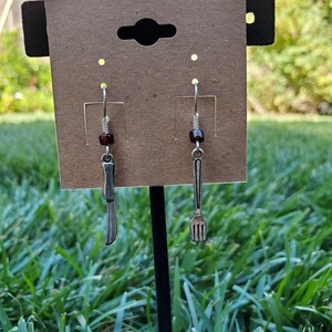May include: A pair of silver earrings with a knife and fork charm. The earrings are hanging from a brown card with a hole in the top.