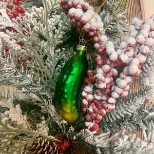 May include: A green glass pickle Christmas ornament with gold accents hangs from a gold string. The ornament is surrounded by a festive arrangement of frosted evergreen branches, red berries, and pinecones.
