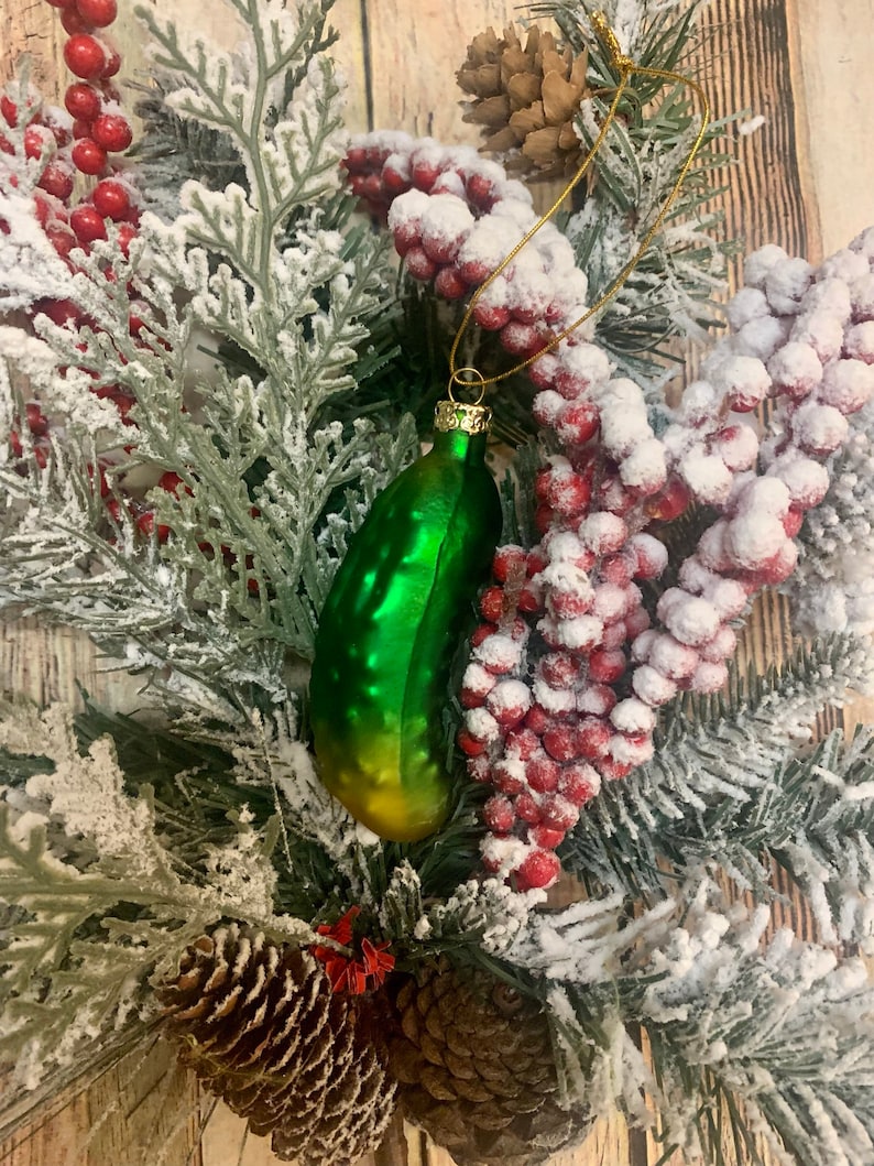 May include: A green glass pickle Christmas ornament with gold accents hangs from a gold string. The ornament is surrounded by a festive arrangement of frosted evergreen branches, red berries, and pinecones.