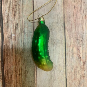 May include: A green glass pickle Christmas ornament with a gold-colored metal loop for hanging. The pickle has a textured surface and a gradient color, transitioning from green to yellow. The ornament is designed to resemble a pickle.
