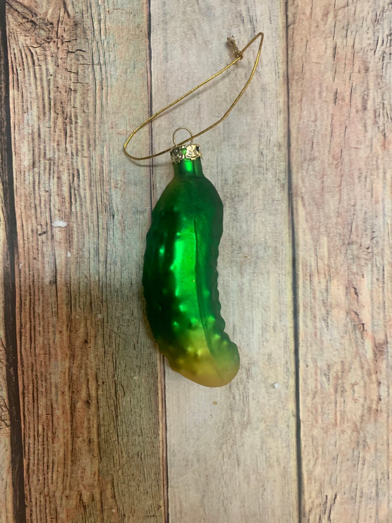 May include: A green glass pickle Christmas ornament with a gold-colored metal loop for hanging. The pickle has a textured surface and a gradient color, transitioning from green to yellow. The ornament is designed to resemble a pickle.