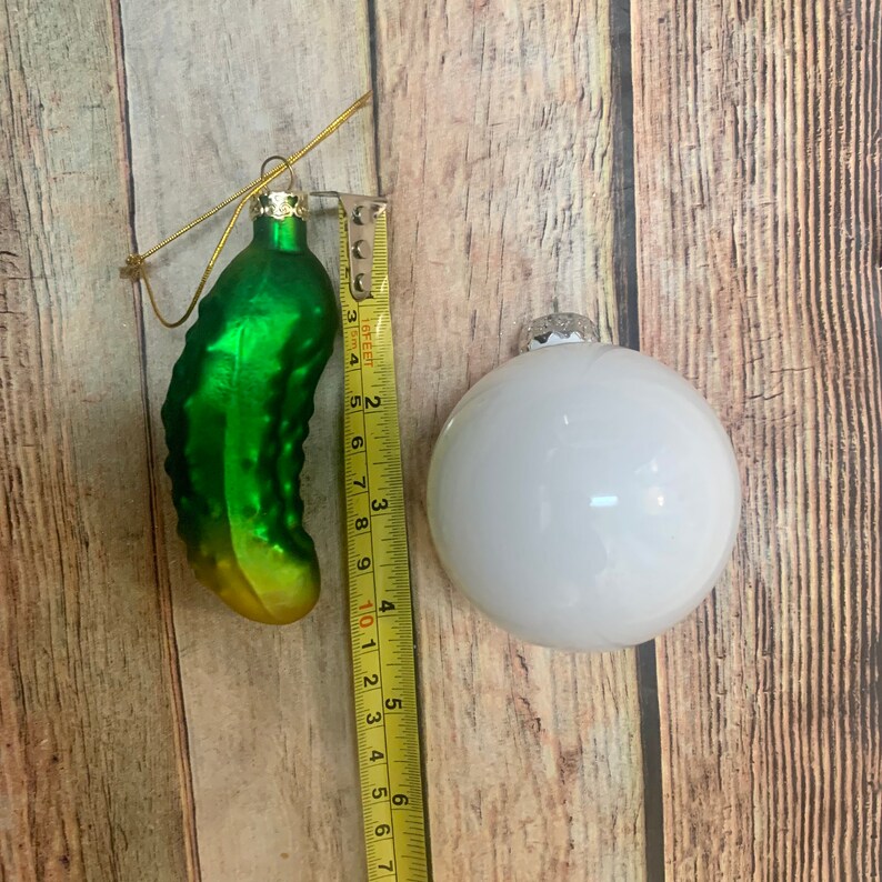 May include: A green pickle-shaped Christmas ornament and a white round ornament. The pickle ornament is approximately 3 inches long. Both ornaments have a gold string for hanging. The ornaments are on a wooden surface.